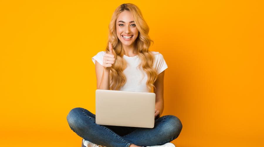 Woman smiling with thumbs up sitting with a laptop against a yellow background